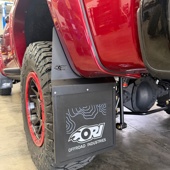 A close-up of a red off-road truck's rear wheel and fender area, showing a black rectangular mudguard with a white ORI Offroad Industries logo. The truck is parked in a garage.